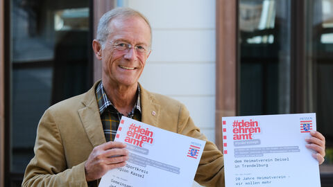 Übergabe Förderbescheide Ehrenamt Kassel Staatsminister Wintermeyer hält zwei Förderbescheide "#deinehrenamt" für Projekte im Landkreis Kassel in die Kamera und lächelt.