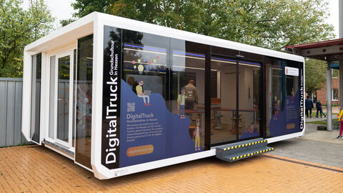 Ein Container-Anhänger aus Glas mit der Aufschrift "DigitalTruck - Grundschultour in Hessen" steht auf einem Schulgelände.