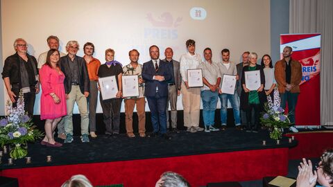 Eine Gruppe steht nebeneinander auf einer Bühne vor Publikum.