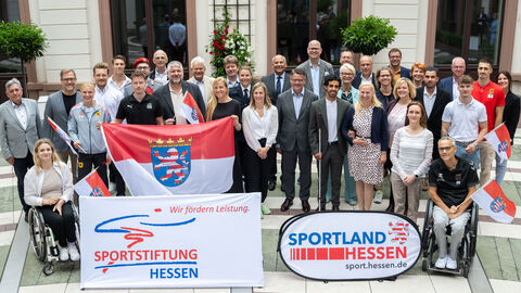 Verabschiedung der Sportlerinnen und Sportler Ministerpräsident Rhein und Sportministerin Stolz stehen inmitten einer Gruppe Sportler, die teilweise Hessen-Flaggen in Händen halten.